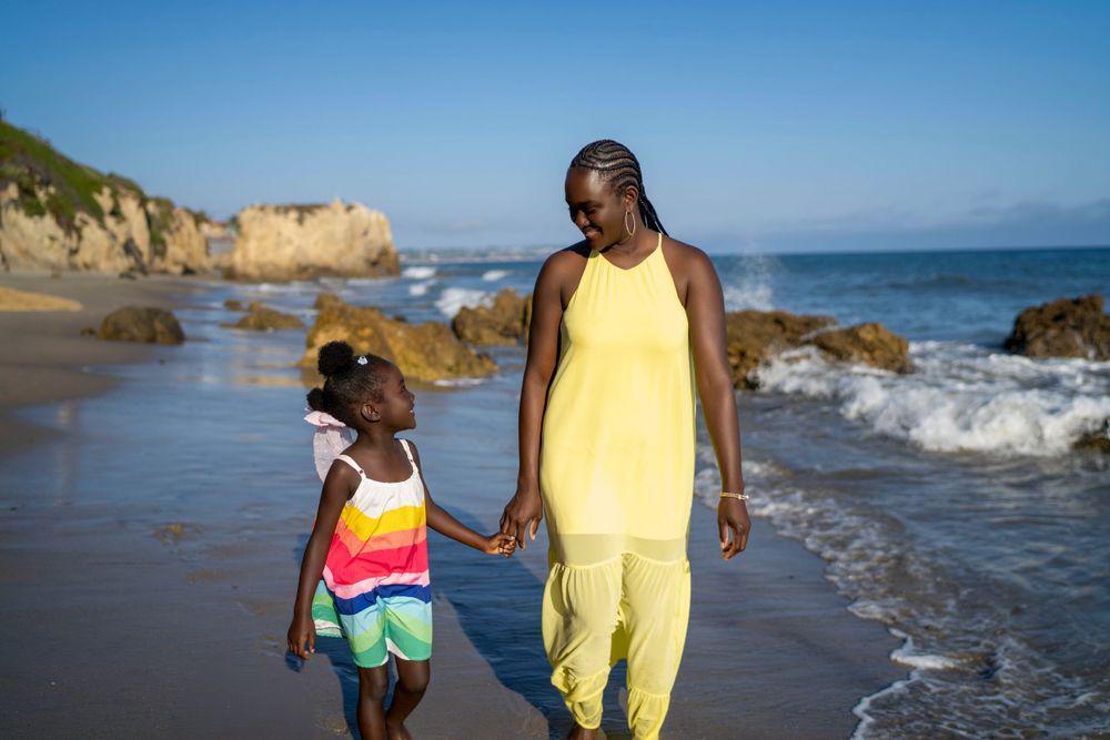 Mom and daughter walking hand in hand on scenic beach with rocky coastline.