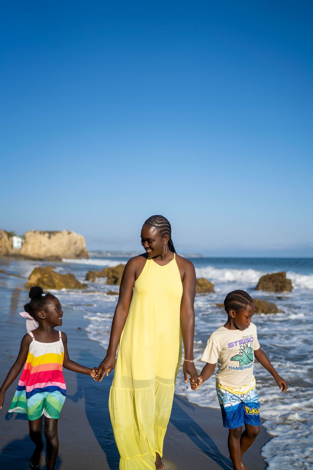 Mom in yellow dress walking with children on sunny beach with coastal rocks in background.