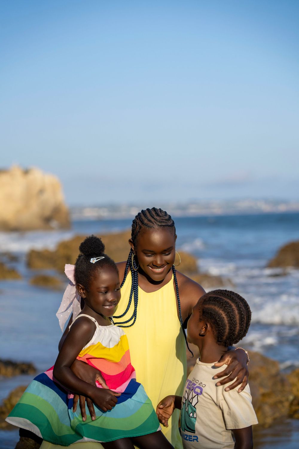 Family enjoying a sunny day at the beach with ocean and rocks in background.