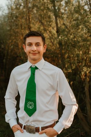 Young person wearing white shirt and green 4-H tie stands against natural wooded background.