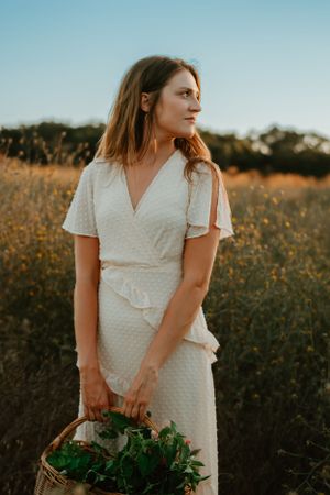 Woman in white dotted dress stands in golden wildflower field at sunset.