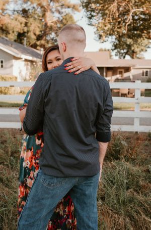 Couple embracing in casual attire against white fence at sunset.