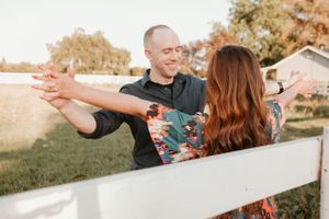 Playful couple leaning on white fence during outdoor countryside photoshoot.
