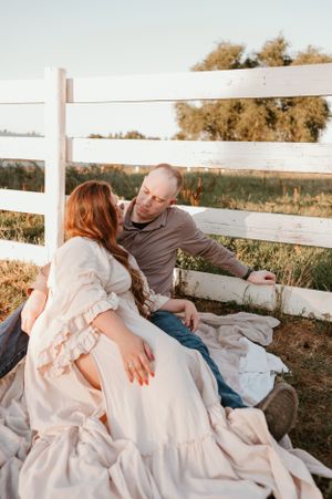 A couple lounges on a white blanket next to a rustic fence during sunset.