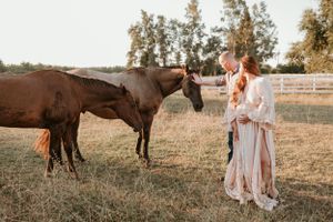 Woman in flowing dress stands near brown horse in pastoral farm setting.