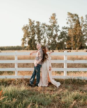 A couple sits by a white fence in a field during golden hour.