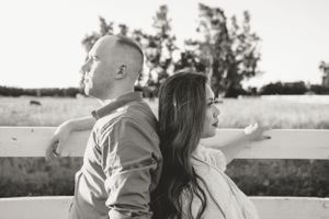 Black and white portrait of couple leaning back-to-back against white fence.