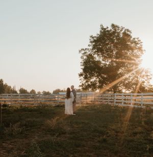 Silhouetted couple stands in field with sunburst through tree at sunset.