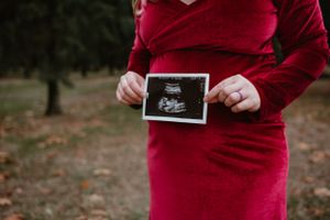 Person in red dress holding ultrasound photos against autumn backdrop.