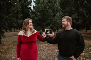 Couple in red and black sharing pregnancy announcement photos in forest setting.