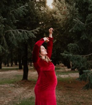 Person in a flowing red velvet dress poses gracefully in a wooded setting.