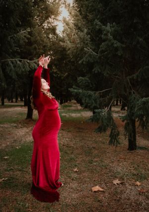 Figure in a dramatic red dress poses expressively against a backdrop of evergreen trees.