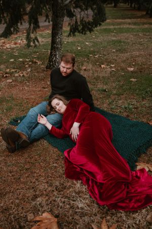 Couple rests on a dark blanket surrounded by fallen leaves in a forest setting.