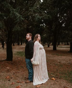 A couple in white and denim walks through a forest during a maternity photo shoot.