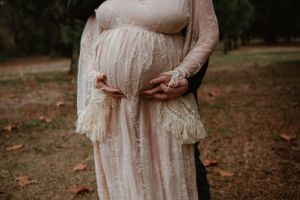 A couple poses for maternity photos wearing a white lace dress and casual attire in a forest.