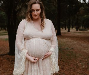 A romantic maternity photo session in a wooded park with a couple wearing a white lace dress and black sweater.