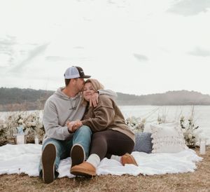 Two people lay on a white blanket during an intimate lakeside moment.