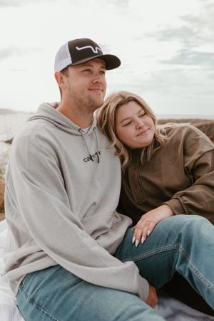 A couple in casual attire shares a tender moment on a coastal hillside.