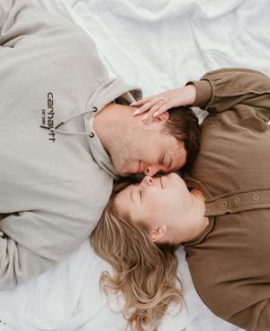 Two people in casual attire sharing a tender moment while lying on white fabric outdoors.