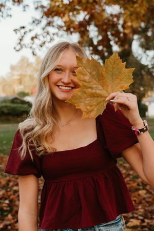 Person in burgundy dress holds large autumn leaf against fall foliage background.