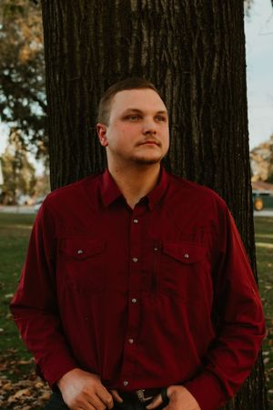 Person in burgundy button-up shirt poses against dark tree trunk background.