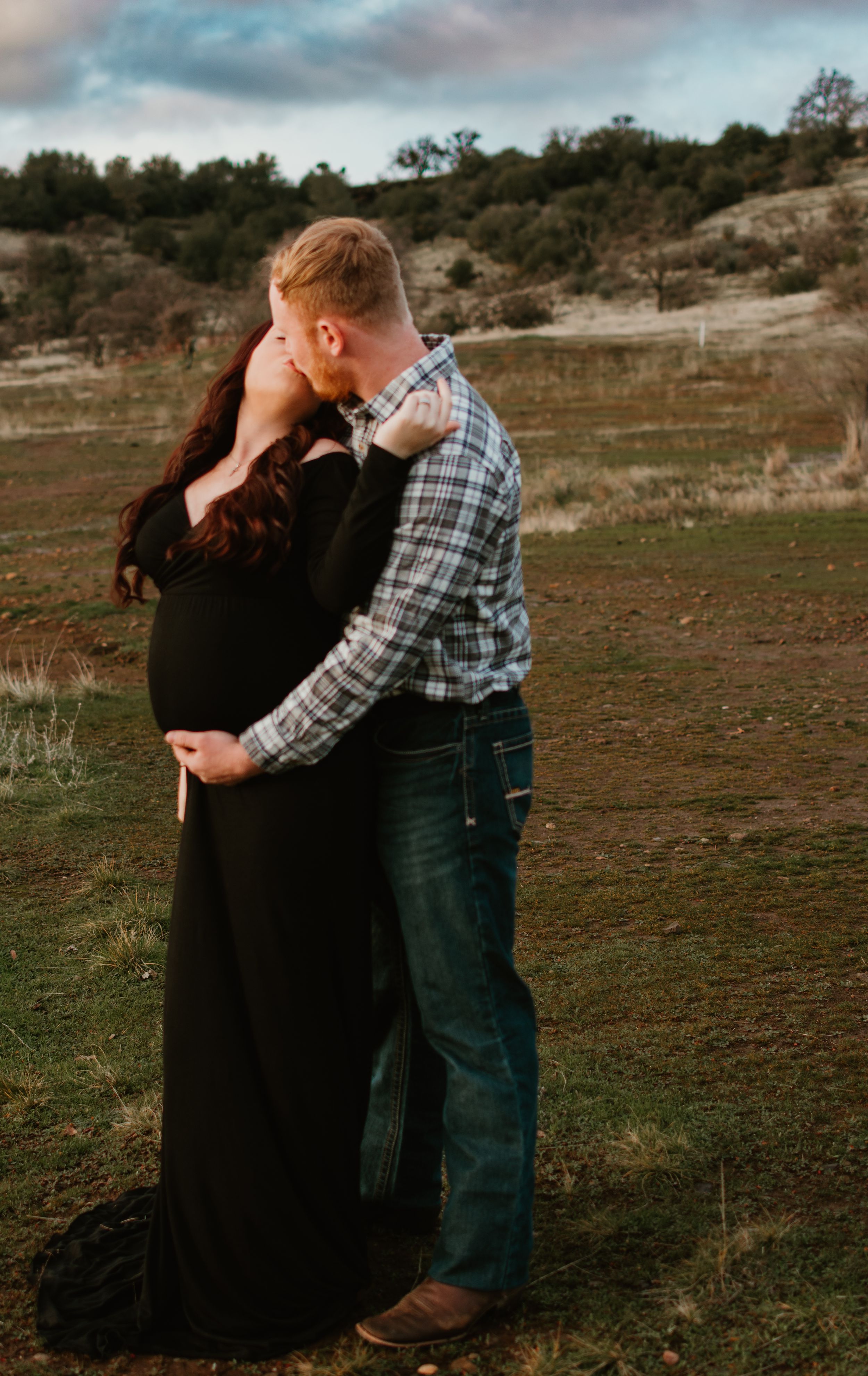 A couple embraces tenderly in a field during sunset while wearing casual attire.