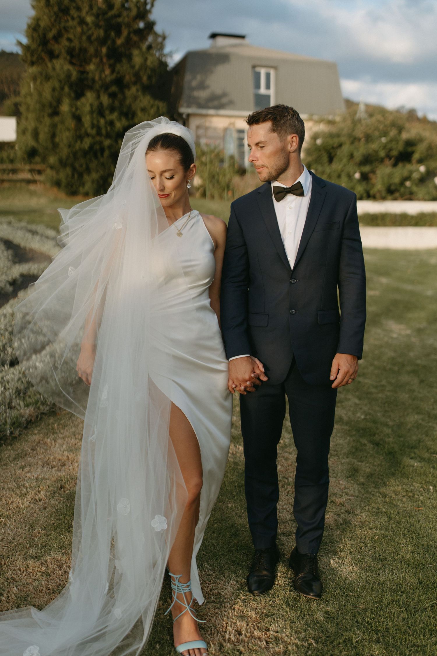 Wedding couple walking on grassy field with bride's veil flowing in the wind.