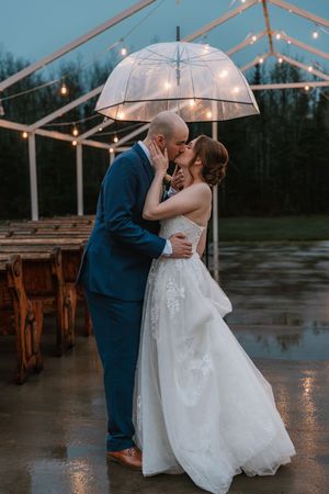Newlyweds sharing a romantic kiss under a clear bubble umbrella on a rainy evening outside a lit tent.