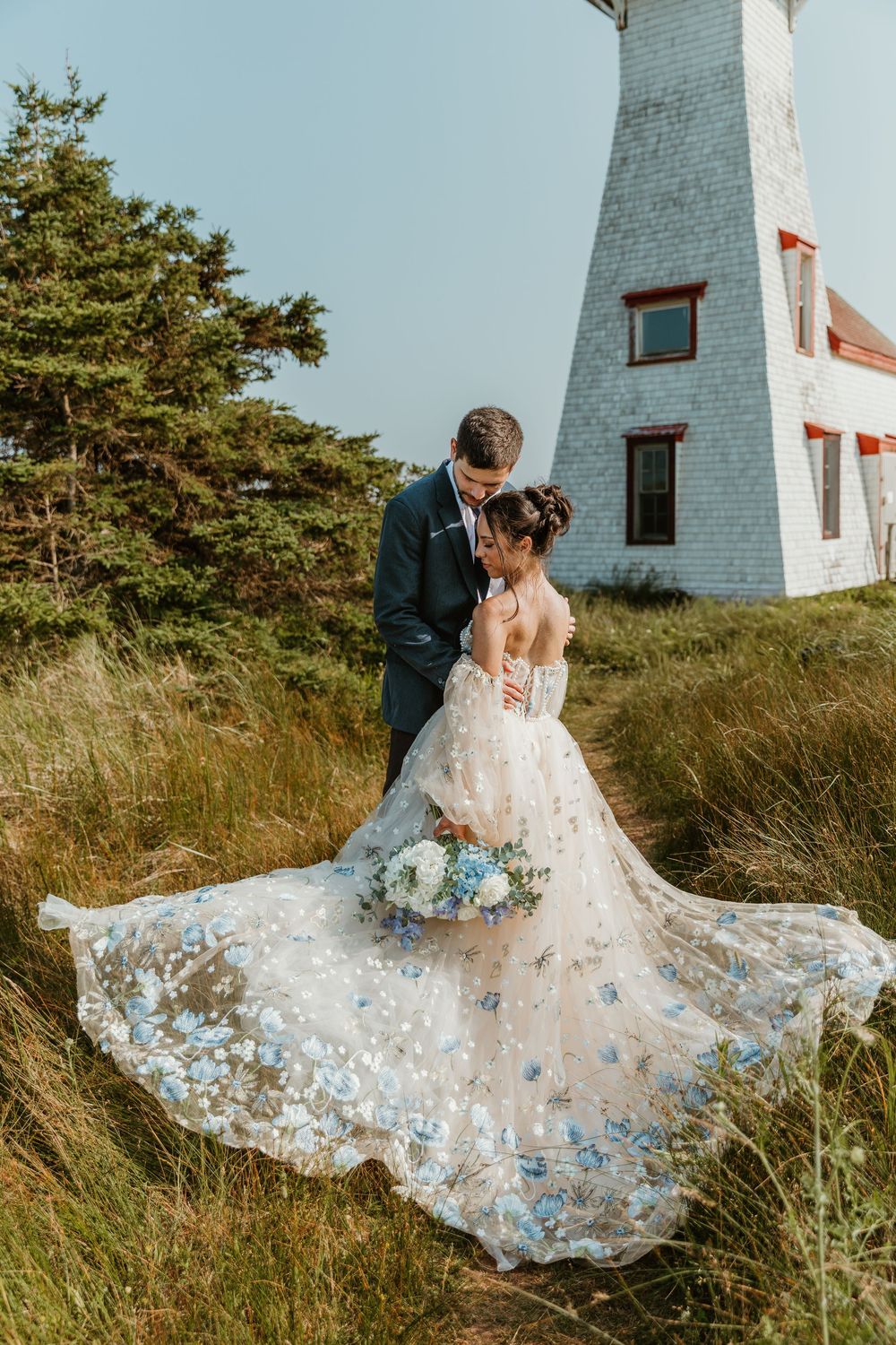 Bride in stunning blue floral gown with flowing train embraces groom beside a classic white lighthouse on PEI