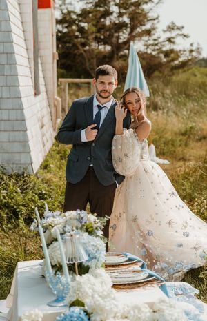 Couple posing beside an elegant outdoor tablescape with blue hydrangeas and a floral wedding gown on Prince Edward Island