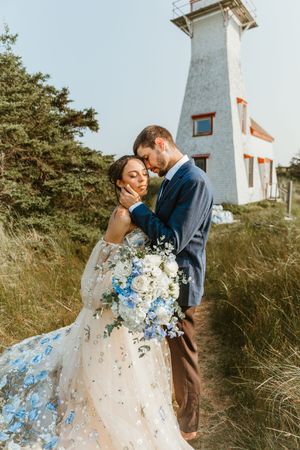 Groom tenderly embracing bride in a floral blue wedding gown near a lighthouse in coastal grass fields on PEI