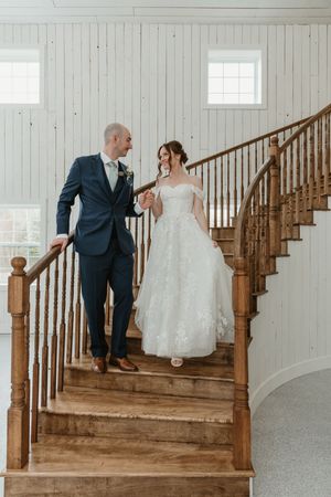 Bride and groom holding hands while descending a wooden staircase in a bright white shiplap interior.