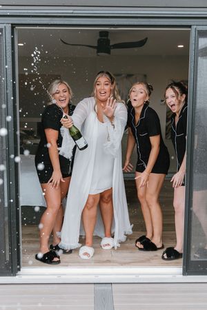Bride in white robe popping champagne with bridesmaids in black pajamas celebrating at a glass doorway.