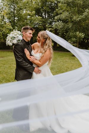 Bride and groom embracing outdoors with flowing white veil billowing in the wind against a lush green backdrop.