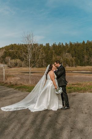 Bride and groom kissing on a rural road with a long cathedral veil and forest backdrop under blue sky in Saint Martins NB