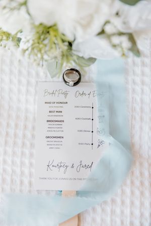 Elegant acrylic wedding program card with rings resting on top, surrounded by white flowers and blue ribbon.