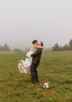 Groom lifting bride in a lace wedding dress for a romantic kiss in a foggy green meadow.