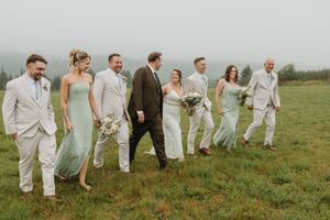 Wedding party in sage green bridesmaid dresses and tan suits walking through a misty green field together.