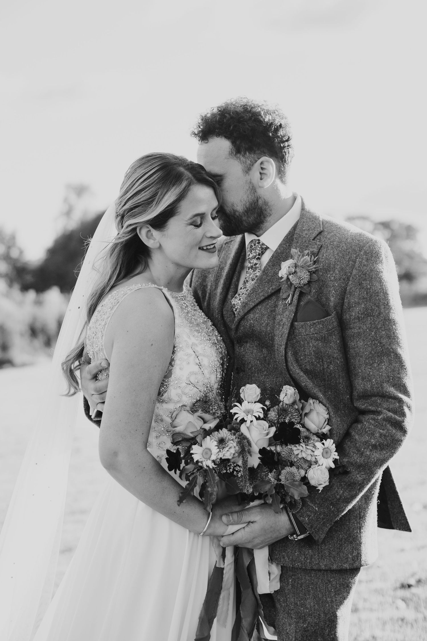 A tender black and white wedding photograph showing a romantic moment as newlyweds embrace while holding a floral bouquet.