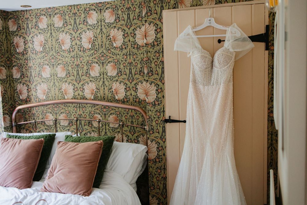 Elegant white lace wedding dress hangs beside a bed with floral wallpaper and pink velvet pillows in a cozy bedroom.