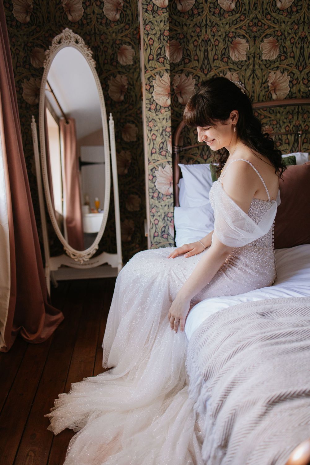 A bride in a delicate lace wedding gown sits on a bed beside an ornate mirror in a room with floral wallpaper.
