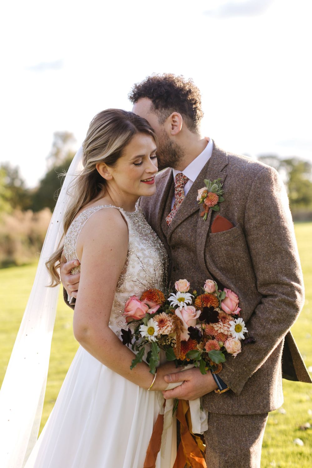 Two newlyweds share an intimate moment outdoors with a lush fall bouquet and brown tweed suit details.