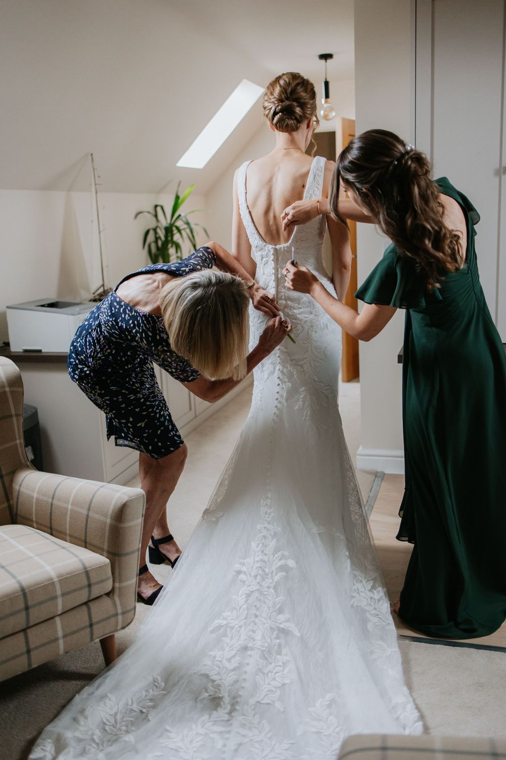 Two women helping to fasten and adjust an elegant lace wedding dress in a bright room with a skylight.