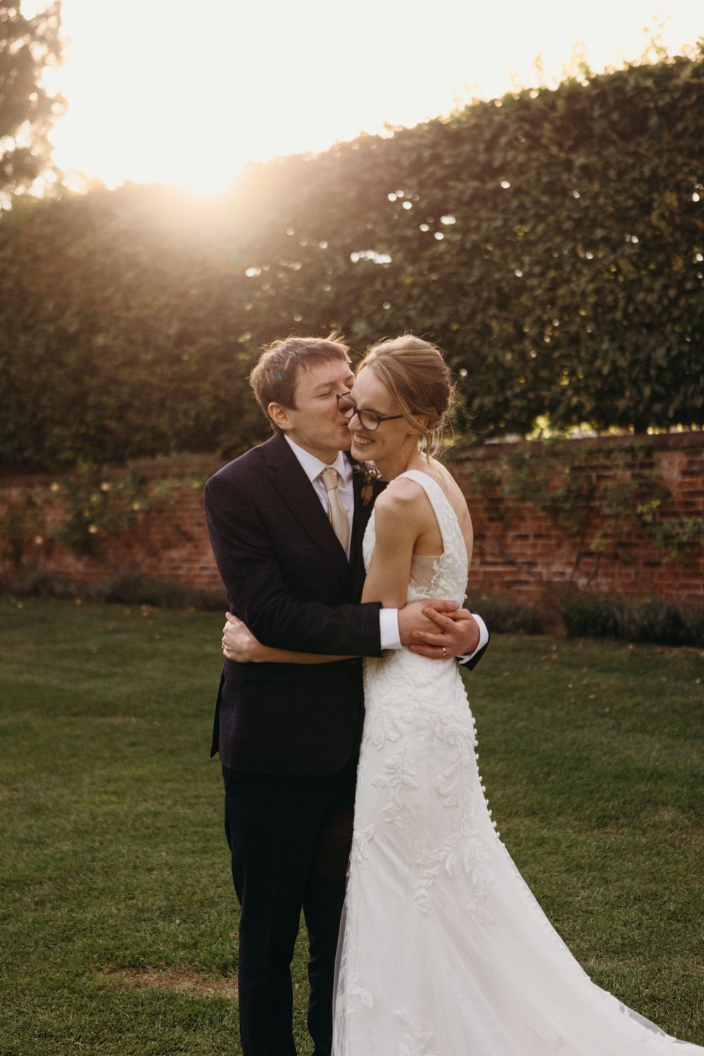 Romantic golden hour wedding photo of couple embracing outdoors with sunlight and garden wall in background.