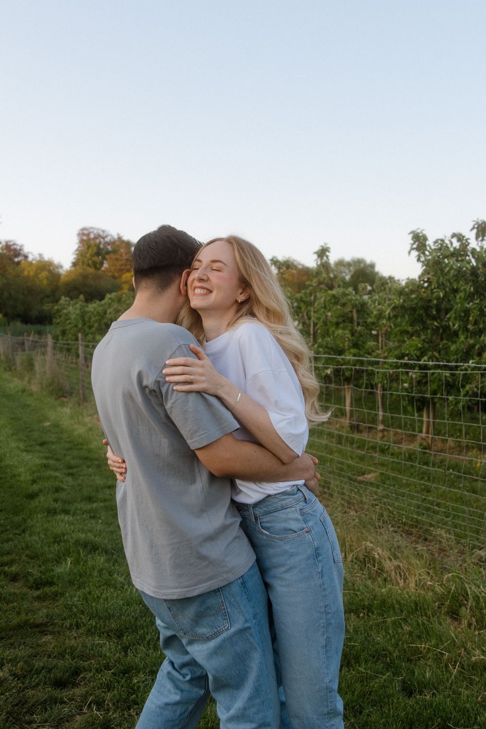 A couple embracing and laughing together in a grassy field with wooden fencing in the background at sunset.