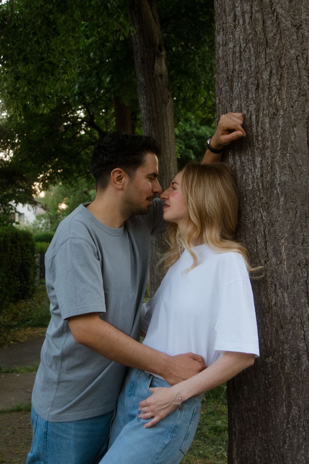 A romantic couple in casual clothing share an intimate moment while leaning against a large tree trunk in a park setting.