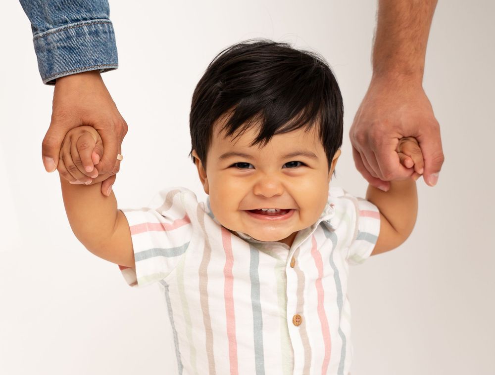A small child wearing a striped shirt smiles while holding hands with adults.