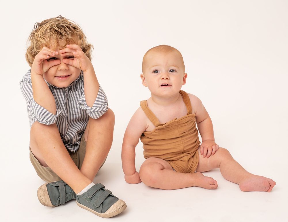 Two young children sit together on a white backdrop wearing casual clothes.