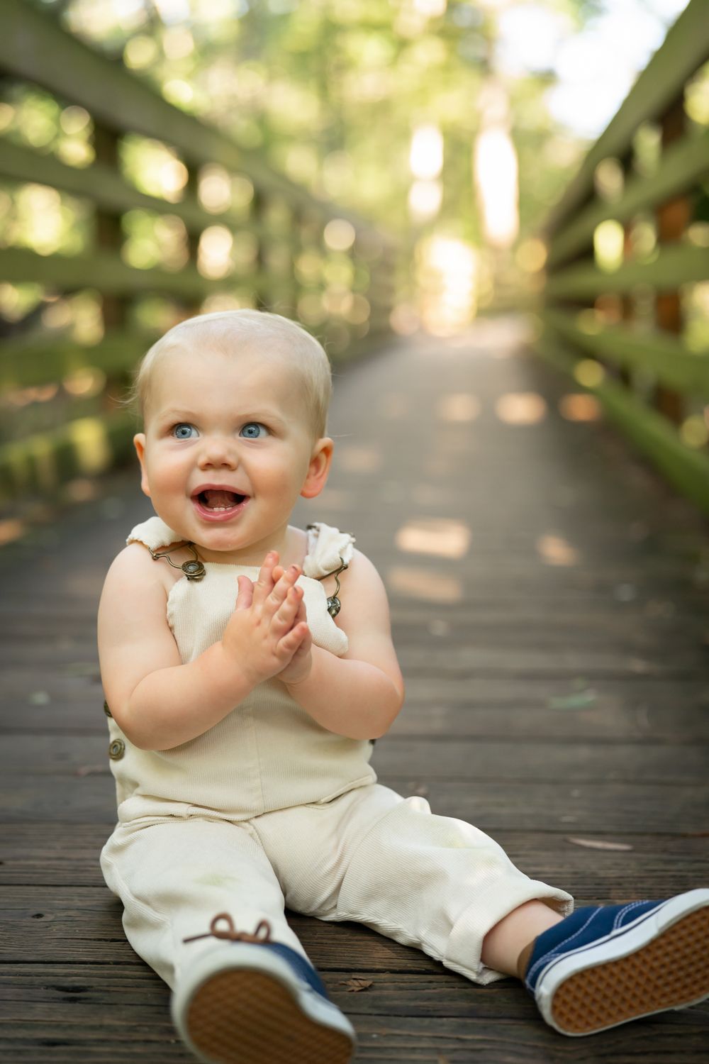 A happy baby in cream-colored overalls sits on a wooden boardwalk surrounded by greenery.