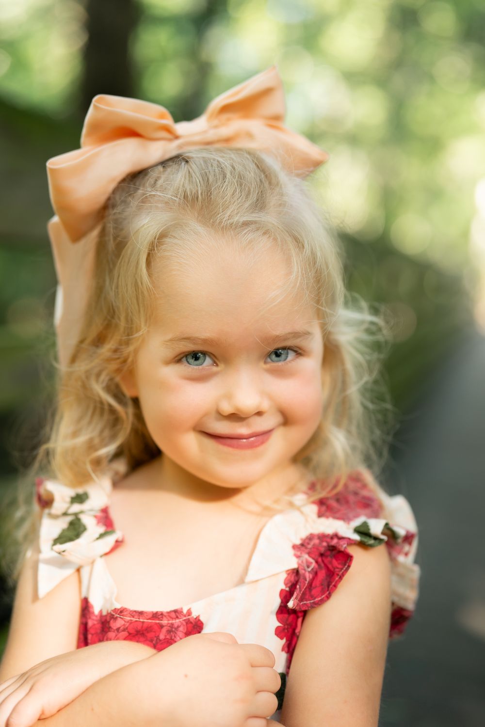 A child in a floral dress with a peach bow smiles in an outdoor setting.